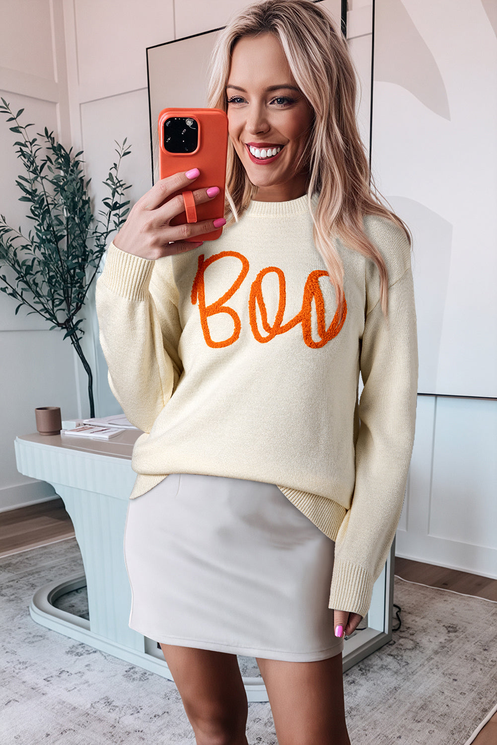 Apricot Halloween Boo Drop Shoulder Knit Sweater with Boo in orange and a beige skirt. A potted plant and decorative table complete the stylish scene.