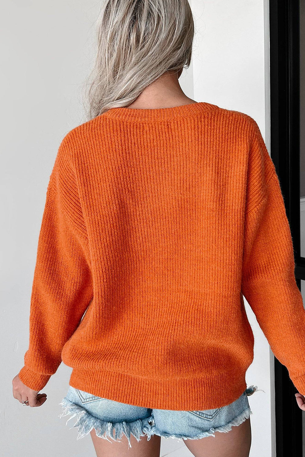 Flamingo Hello Pumpkin Graphic Sweater in orange, paired with light blue distressed denim shorts. The background is plain with a door frame partly visible.