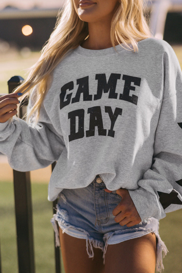 A woman with long blonde hair wears a relaxed fit Black Game Day Graphic Rugby Football Season Sweatshirt and distressed denim shorts, standing outdoors near a fence—perfect for rugby football fans.